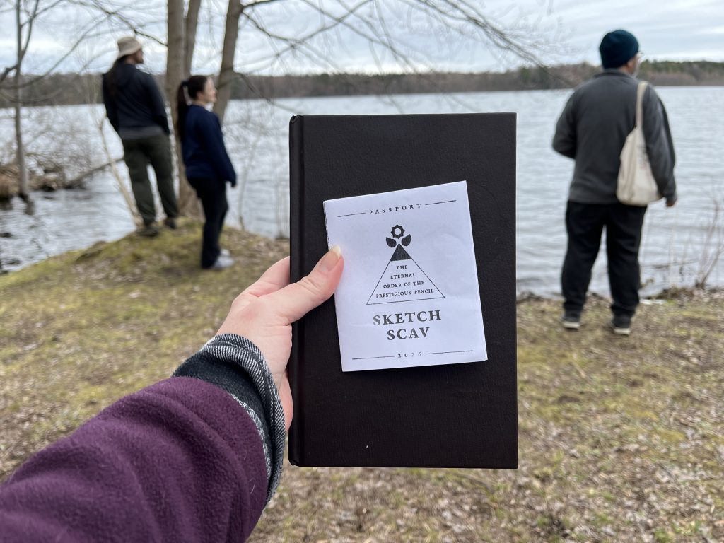 IMG_6840 An outstretched arm and hand hold a sketchbook and an EOPP Sketch Scav passport zine on a backdrop of the outdoors near a lake. In the background, three people look out over the lake.